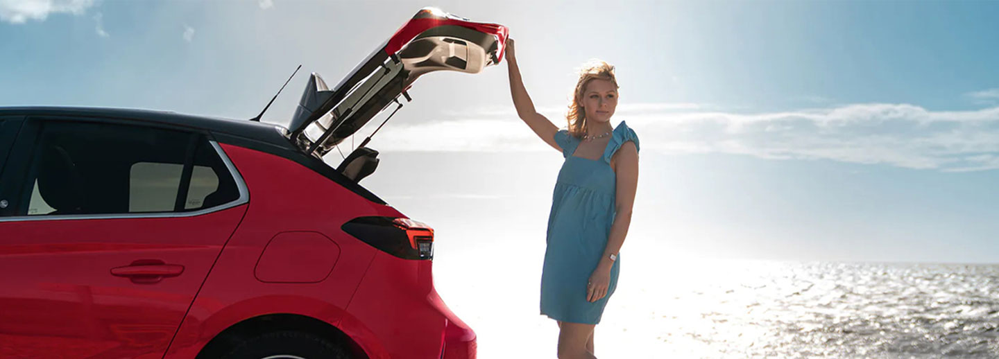 Lady on a beach, standing to the rear of a Vauxhall Corsa with boot open