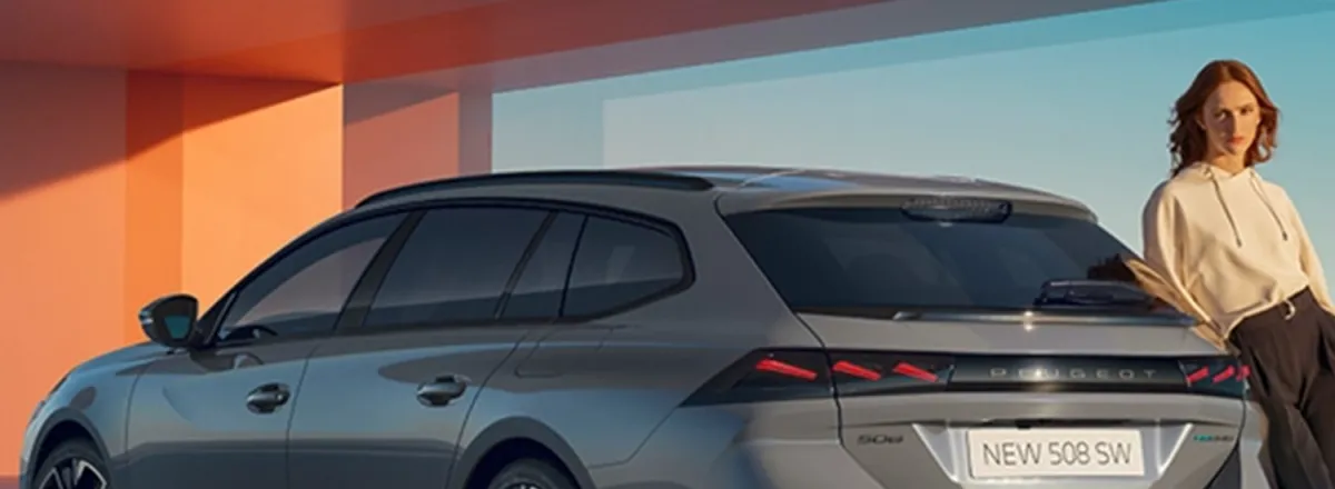 A lady leaning against the side of a cumulus grey Peugeot 508 SW car. The car is parked in front of an orange building structure.