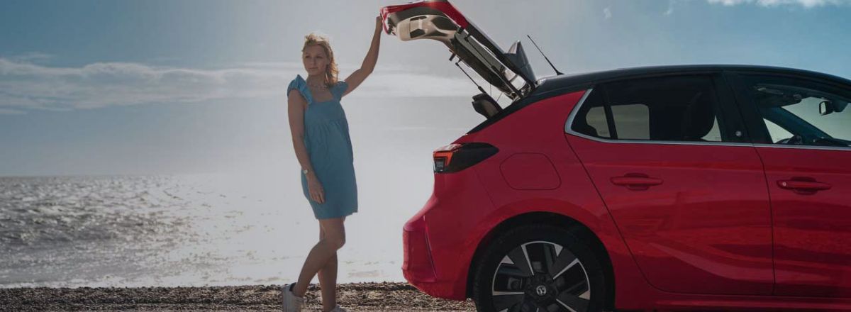 Red Vauxhall Corsa on a pebble beach with lady standing by the boot