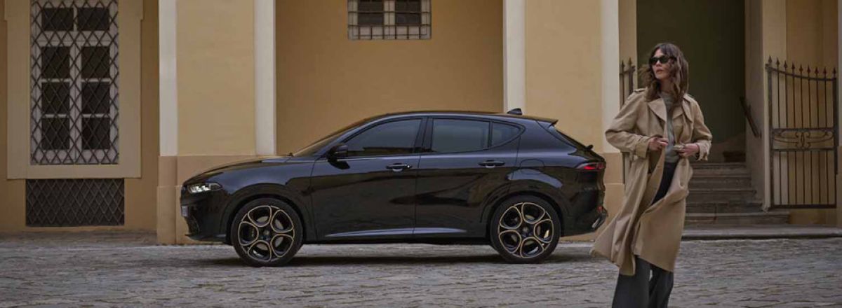 Tonale in Alfa Black parked with the passenger side facing on a cobblestone street in front of a yellow building with arched doorways and windows, with a person in a long beige coat standing in the foreground.
