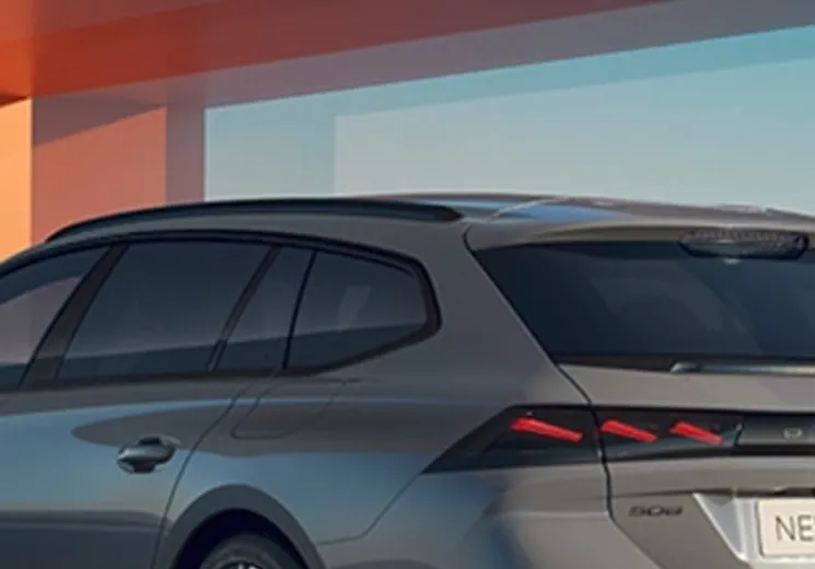 A lady leaning against the side of a cumulus grey Peugeot 508 SW car. The car is parked in front of an orange building structure.