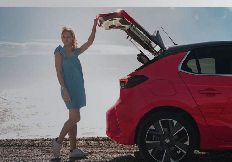 Red Vauxhall Corsa on a pebble beach with lady standing by the boot