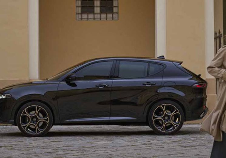 Tonale in Alfa Black parked with the passenger side facing on a cobblestone street in front of a yellow building with arched doorways and windows, with a person in a long beige coat standing in the foreground.