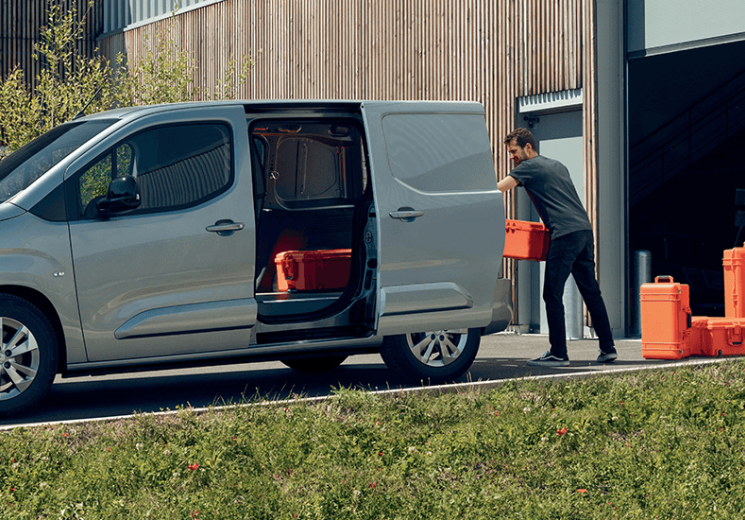 Man wearing a grey shirt loading a silver Peugeot Expert with orange tool boxes. 