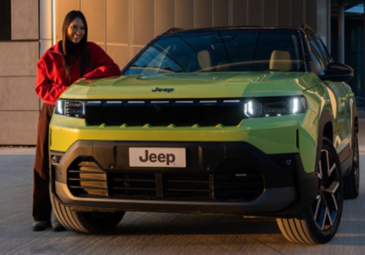 Person standing beside a lime green Jeep Compass Electric parked outside a modern building, viewed from the front of the vehicle.