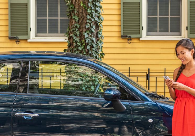 Person standing beside a blue car, using a smartphone outside a residential property.