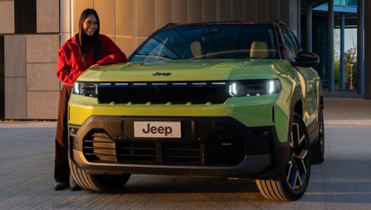 Person standing beside a lime green Jeep Compass Electric parked outside a modern building, viewed from the front of the vehicle.