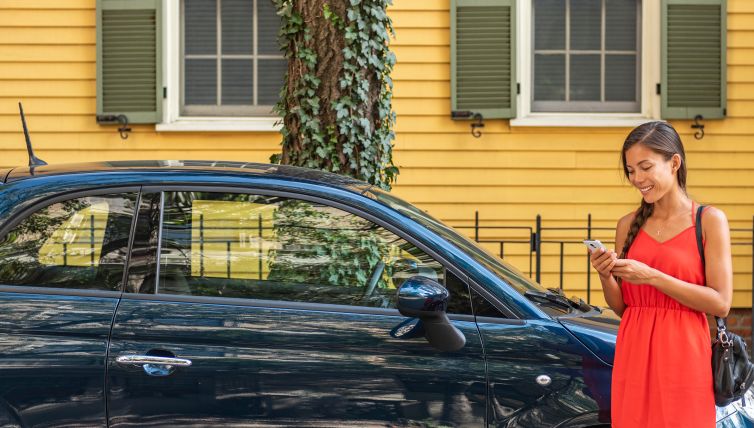 Person standing beside a blue car, using a smartphone outside a residential property.